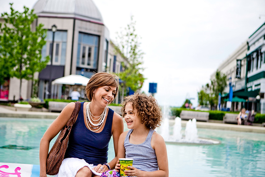 A woman and her daughter sitting on the edge of a fountain outside a shopping center