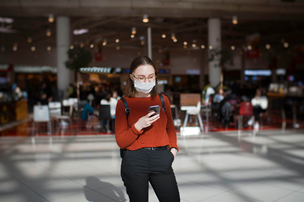 Woman in a red sweater wearing a mask and looking at her phone in a mall food court
