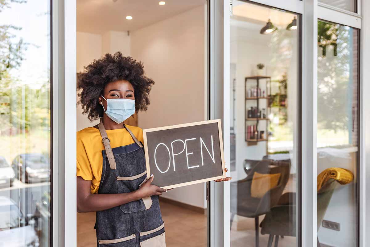 A woman wearing a mask and holding a sign that says "Open" in the doorway of her salon